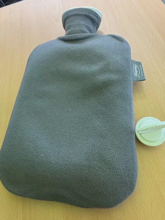 회색 플리스 커버를 씌운 파쉬 보온 물주머니가 책상 위에 놓여 있고, 옆에 연두색 마개가 분리되어 있다